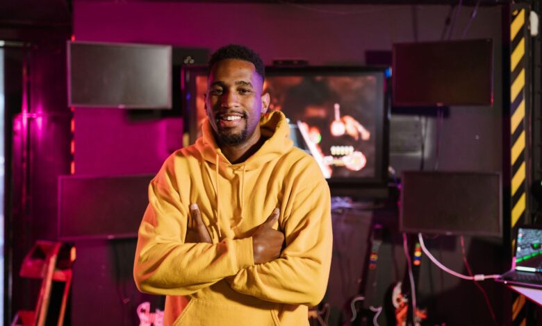 Smiling gamer in a yellow hoodie stands confidently in an e-sports studio with vibrant lighting.