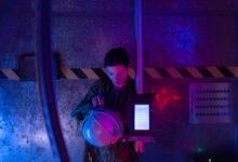 A female astronaut in a spacesuit examines her helmet in a dimly lit spaceship interior.