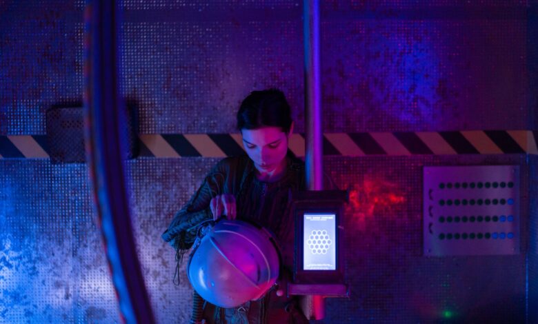 A female astronaut in a spacesuit examines her helmet in a dimly lit spaceship interior.