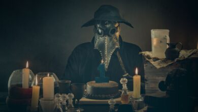 A masked figure resembling a plague doctor sits with candles and a cake in a dimly lit room.