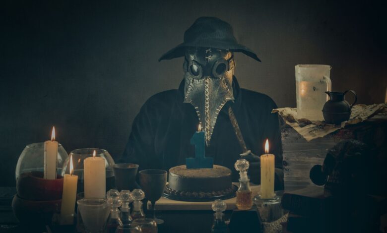A masked figure resembling a plague doctor sits with candles and a cake in a dimly lit room.