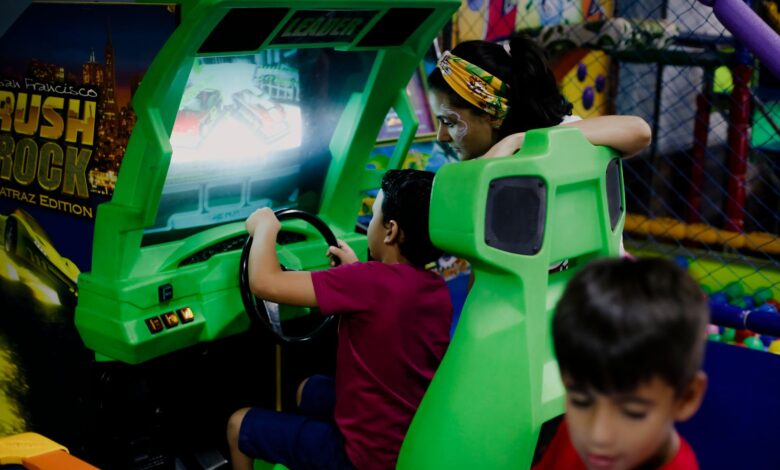 Unrecognizable child having fun with bright car racing simulator while turning steering wheel near serious ethnic woman and funny brother in shopping center near playground