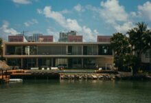 A modern waterfront mansion featuring unique architecture, surrounded by palm trees under a clear blue sky.