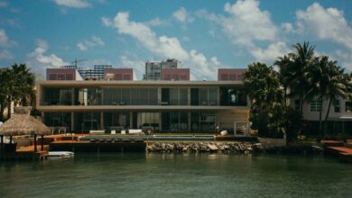 A modern waterfront mansion featuring unique architecture, surrounded by palm trees under a clear blue sky.