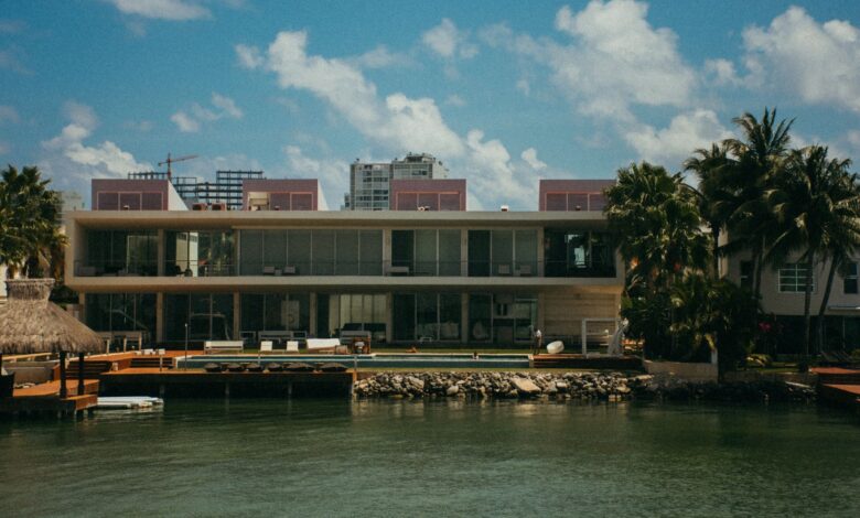 A modern waterfront mansion featuring unique architecture, surrounded by palm trees under a clear blue sky.