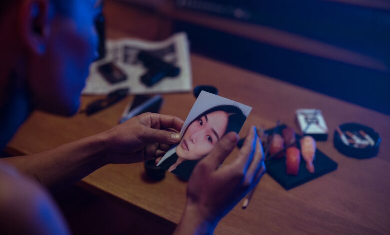 A person holds a photo in a dimly lit room, surrounded by objects suggesting mystery.