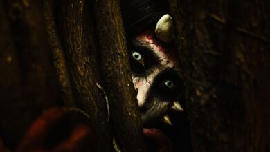 A person with devilish makeup peers through tree branches, creating an eerie Halloween vibe.