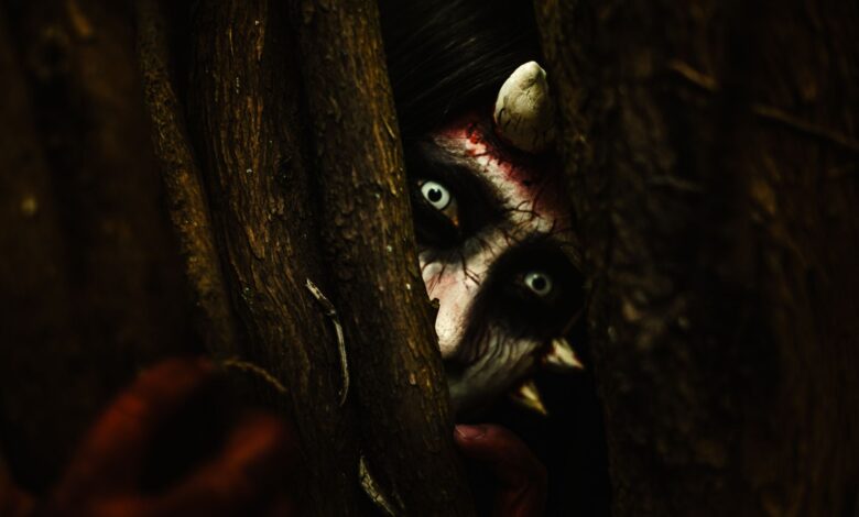 A person with devilish makeup peers through tree branches, creating an eerie Halloween vibe.
