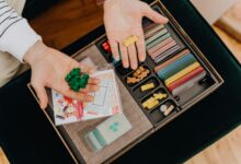 Colorful board game components being held in hands above a game box, top view.