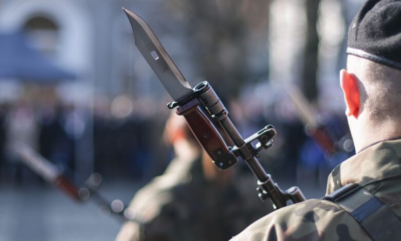 Military personnel holding rifle with bayonet during an outdoor ceremonial event.