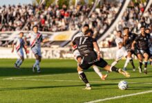 A dynamic football match with players in motion on a sunny day, witnessed by a packed stadium.