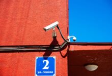 A vivid red wall featuring surveillance cameras and a number sign under a clear blue sky.
