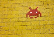 Street art featuring a retro pixelated space invader on a vivid yellow brick wall.