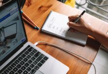 A person writing notes while researching online courses on a laptop in a home setting.