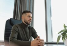 Bearded businessman in turtleneck at work, seated by a bright window.