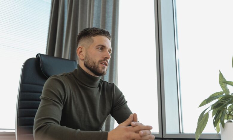 Bearded businessman in turtleneck at work, seated by a bright window.