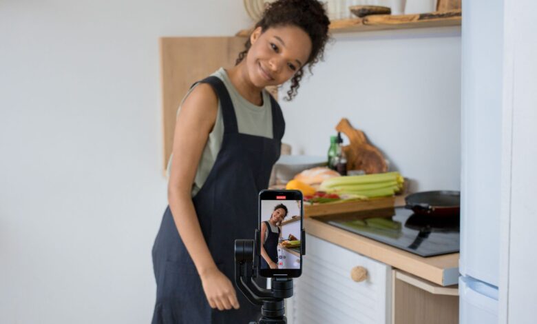 Smiling young woman films a cooking video at home using a smartphone in her kitchen.