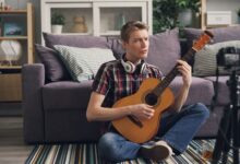 Man sitting cross-legged playing acoustic guitar indoors, streaming online.
