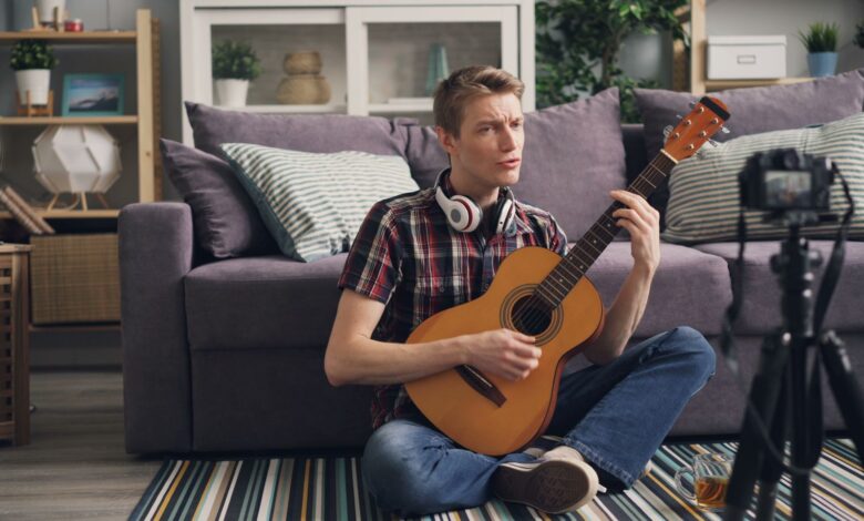 Man sitting cross-legged playing acoustic guitar indoors, streaming online.