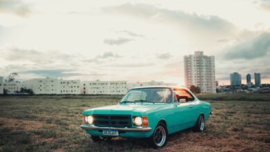 A vintage turquoise car parked in a field with a city skyline backdrop at sunset, capturing urban nostalgia.