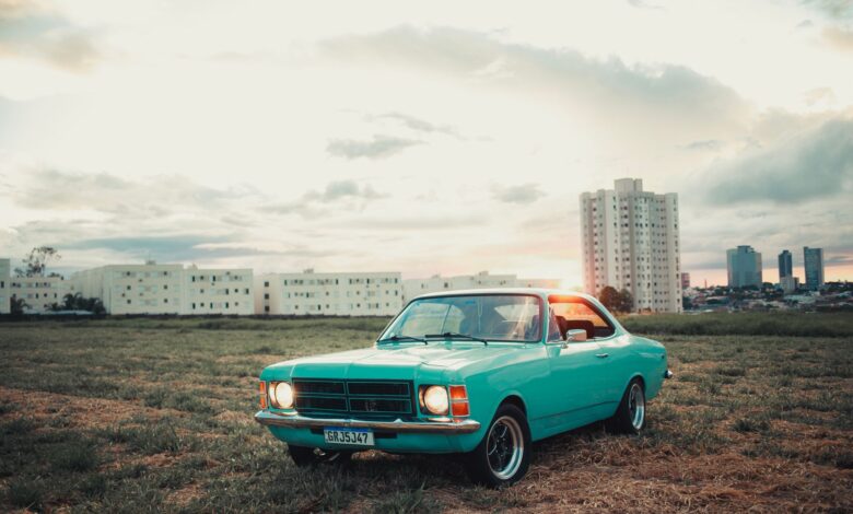 A vintage turquoise car parked in a field with a city skyline backdrop at sunset, capturing urban nostalgia.