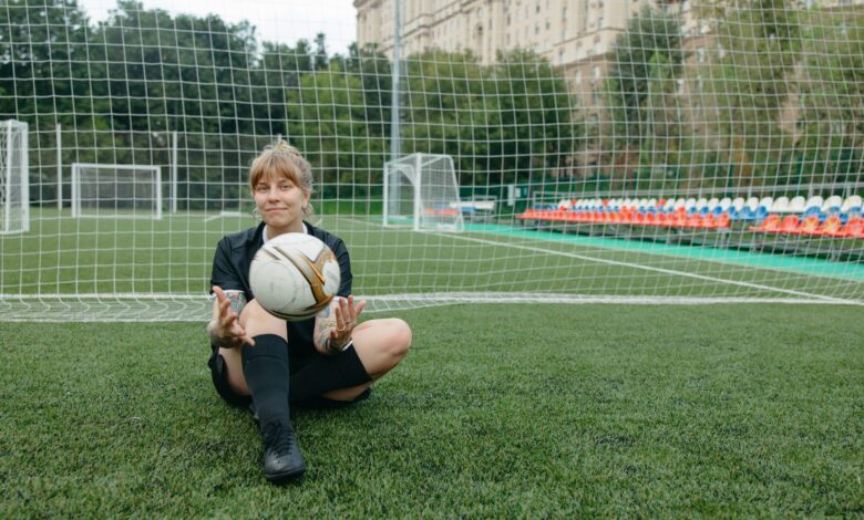 A female soccer player sitting with a ball on a soccer field, enjoying a break.