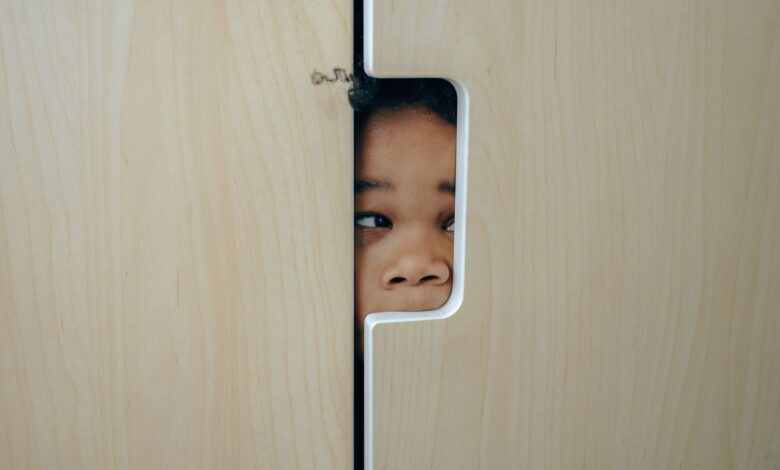 Anonymous funny little ethnic child hiding in wardrobe and peeping through hole while playing at home