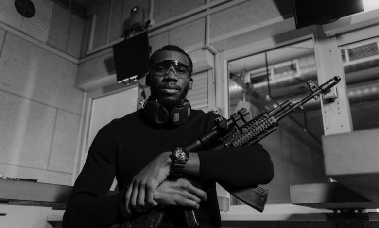 Black and white image of a man with a rifle at a shooting range, wearing safety equipment.