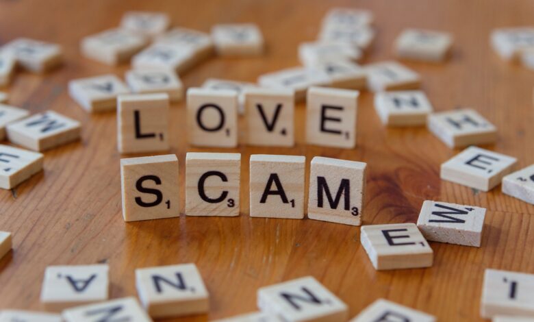 Scrabble tiles form words 'LOVE' and 'SCAM' on a wooden surface, illustrating deceit in relationships.