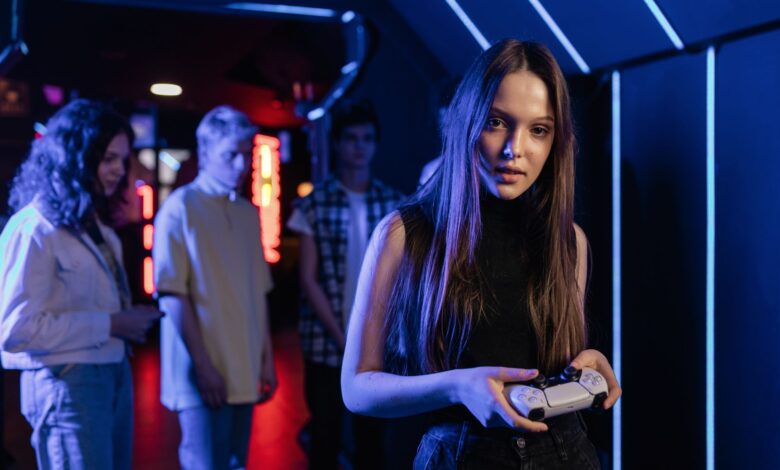 A young woman holding a game controller in a neon-lit arcade, surrounded by friends.