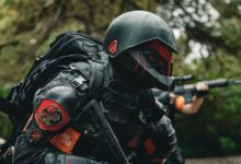 A soldier in advanced tactical armor with a firearm crouches in a forested area during a mission.