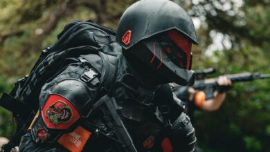 A soldier in advanced tactical armor with a firearm crouches in a forested area during a mission.