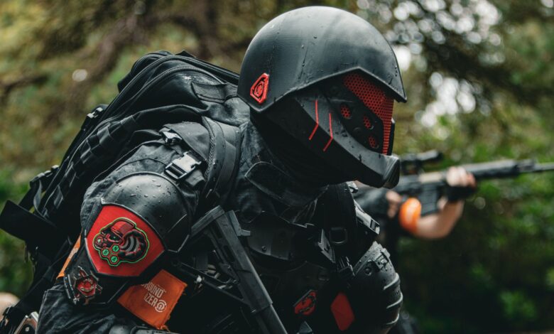 A soldier in advanced tactical armor with a firearm crouches in a forested area during a mission.
