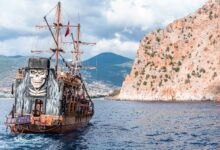 Wooden pirate boat with sailing mast and painted skull floating on rippling sea near rocky mountain against grassy hilly area