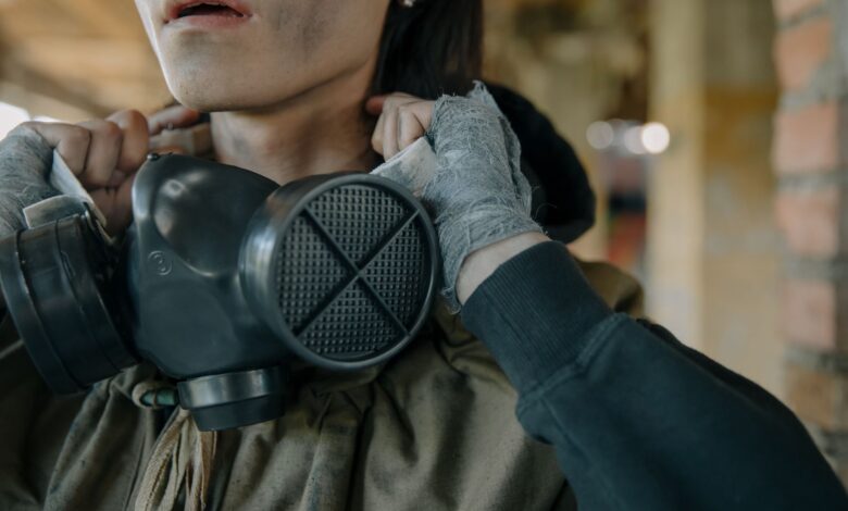 A person with a gas mask and bandaged hands in an urban setting, indicating survival theme.