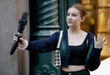 Young woman in casual attire takes a selfie outdoors using a smartphone and stand.