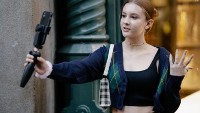 Young woman in casual attire takes a selfie outdoors using a smartphone and stand.