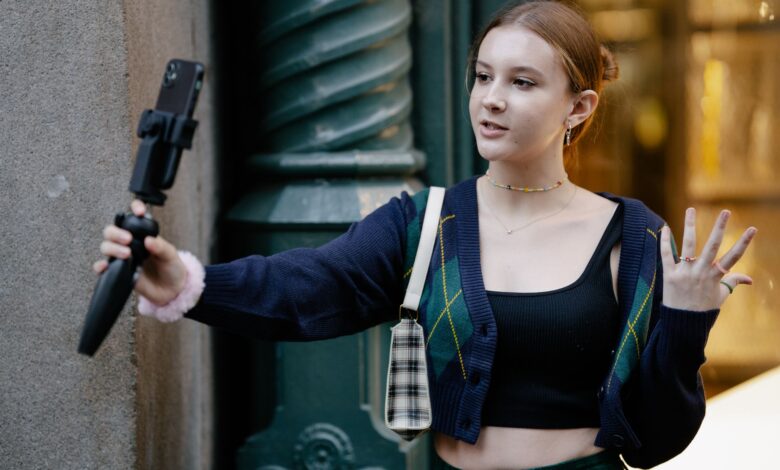 Young woman in casual attire takes a selfie outdoors using a smartphone and stand.