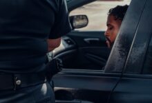 A police officer issuing a ticket to a man seated in a car, captured outdoors in daylight.