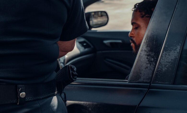 A police officer issuing a ticket to a man seated in a car, captured outdoors in daylight.