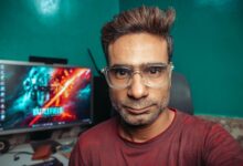 A man with glasses poses for a selfie in his home office, with a computer screen in the background.