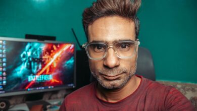 A man with glasses poses for a selfie in his home office, with a computer screen in the background.