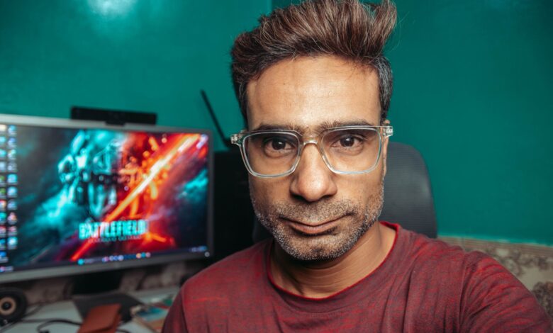 A man with glasses poses for a selfie in his home office, with a computer screen in the background.