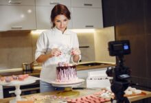 Smiling female pastry chef in uniform decorating cake and looking at lens while standing in front of video camera in modern bakery