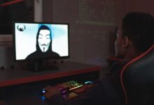 Person using computer in dark room viewing hacker wearing a mask on the screen, symbolizing cyber security threats.