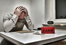 Bald man holding head in despair at desk with cashbox and money. Indoor office stress concept.