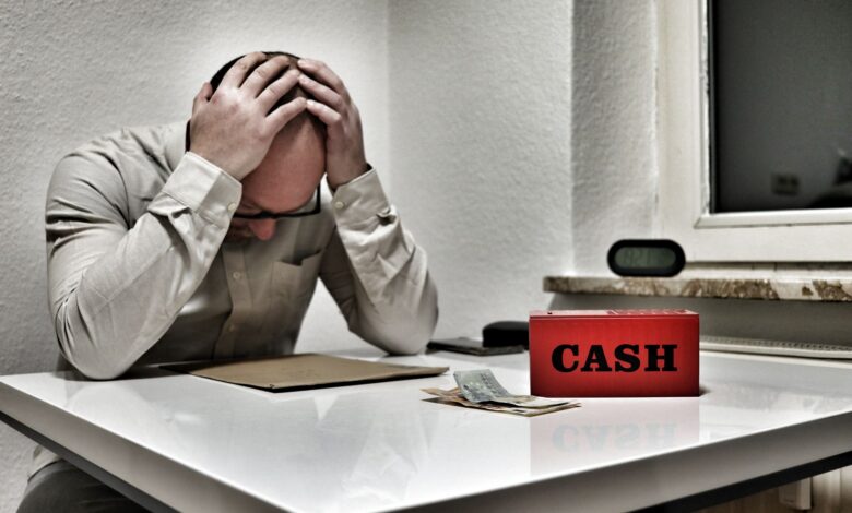 Bald man holding head in despair at desk with cashbox and money. Indoor office stress concept.