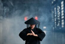 A martial artist in conical hat and black outfit performing a pose in an industrial setting.