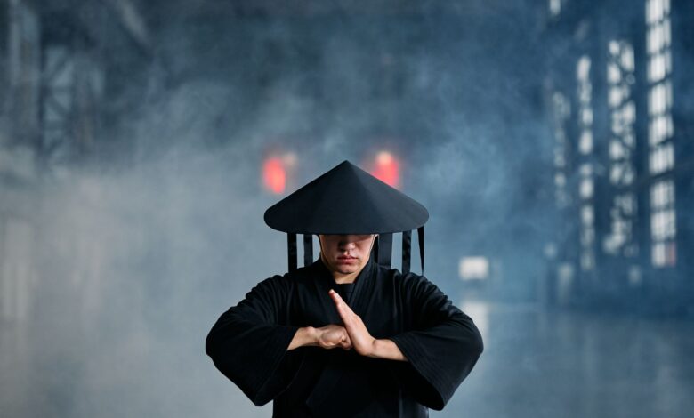 A martial artist in conical hat and black outfit performing a pose in an industrial setting.