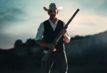 A cowboy in traditional attire holding a shotgun, standing outdoors in San Antonio, Texas.